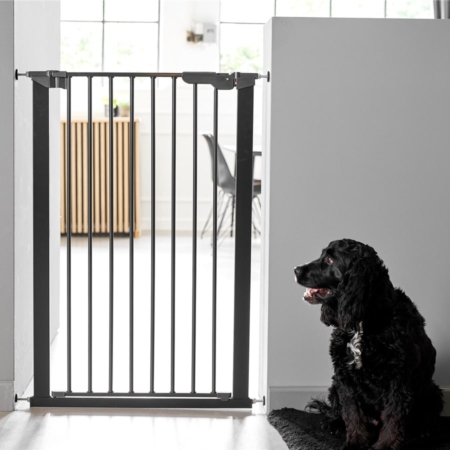 Kutyarács-DogSpace Bonie Extra magas Kutyarács, mely egy ajtó nyilásba van felszerelve. A kutya rács fekete színű, s előtte ül egy fekete színű kutya. A Kutyarács ajtaja be van csukva.