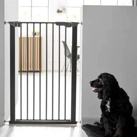 Kutyarács-DogSpace Bonie Extra magas Kutyarács, mely egy ajtó nyilásba van felszerelve. A kutya rács fekete színű, s előtte ül egy fekete színű kutya. A Kutyarács ajtaja be van csukva.