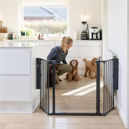 Kutyarács - Dogspace M fekete fém kutyarács. A konyhában van felszerelve. Egyik fele a falhoz van rögzítve a másik pedig a konyhabútorhoz. A kutyarács ajtaja nyitva van és a háttérben egy nő készül megetetni 2 barna színű kutyát.