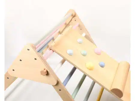 Pikler féle háromszög rámpával. A képen a mászóka látható, melíynek a rudjai pasztel színűek. A rámpa rá van akasztva a háromszögre és rámpán vannak kapszkodók pasztel színben.